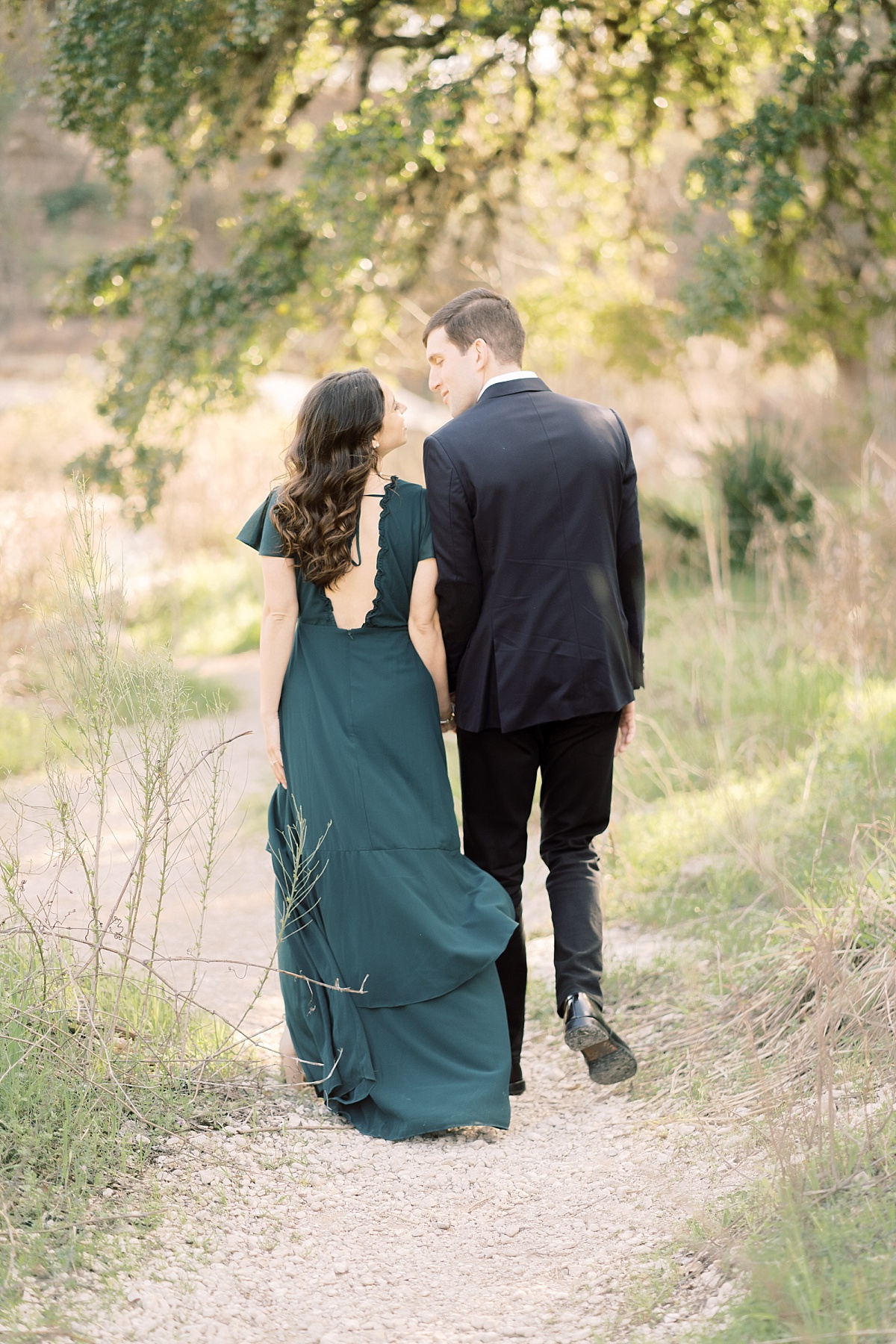 An attractive couple looks lovingly at each other while holding hands and walking away from the camera in a beautiful park.