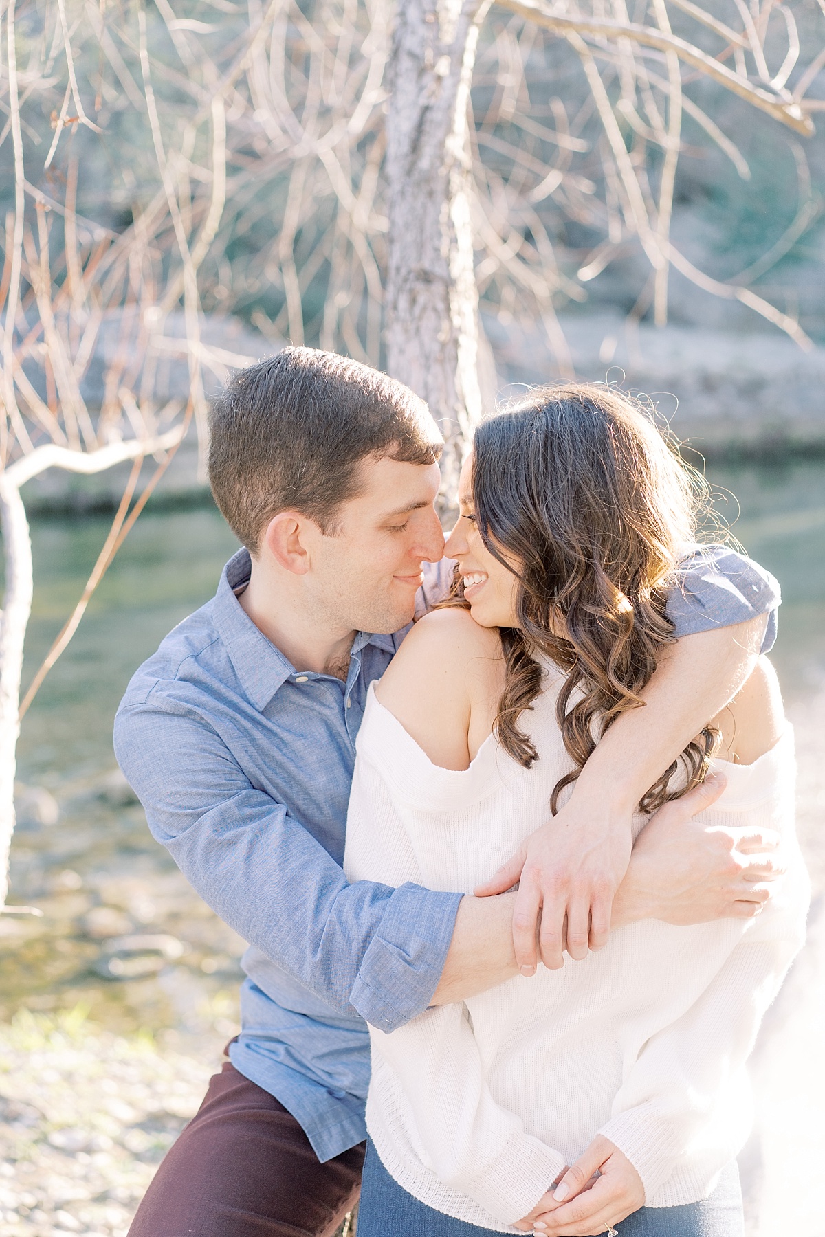 A man in a collared shirt wraps his arms around his beautiful fiance in a bright outdoor setting.