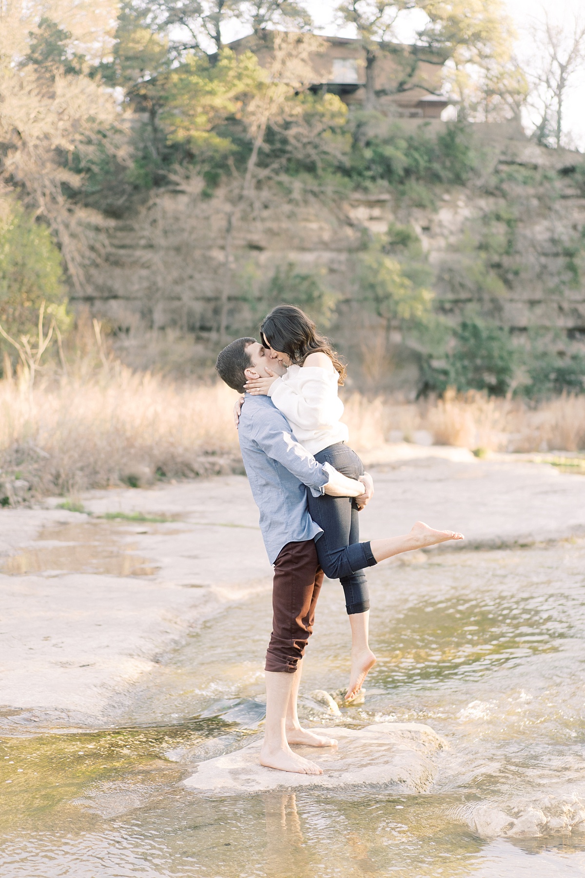 A tall man lifts and kisses his fiance while standing barefoot in the Colorado River at golden hour.