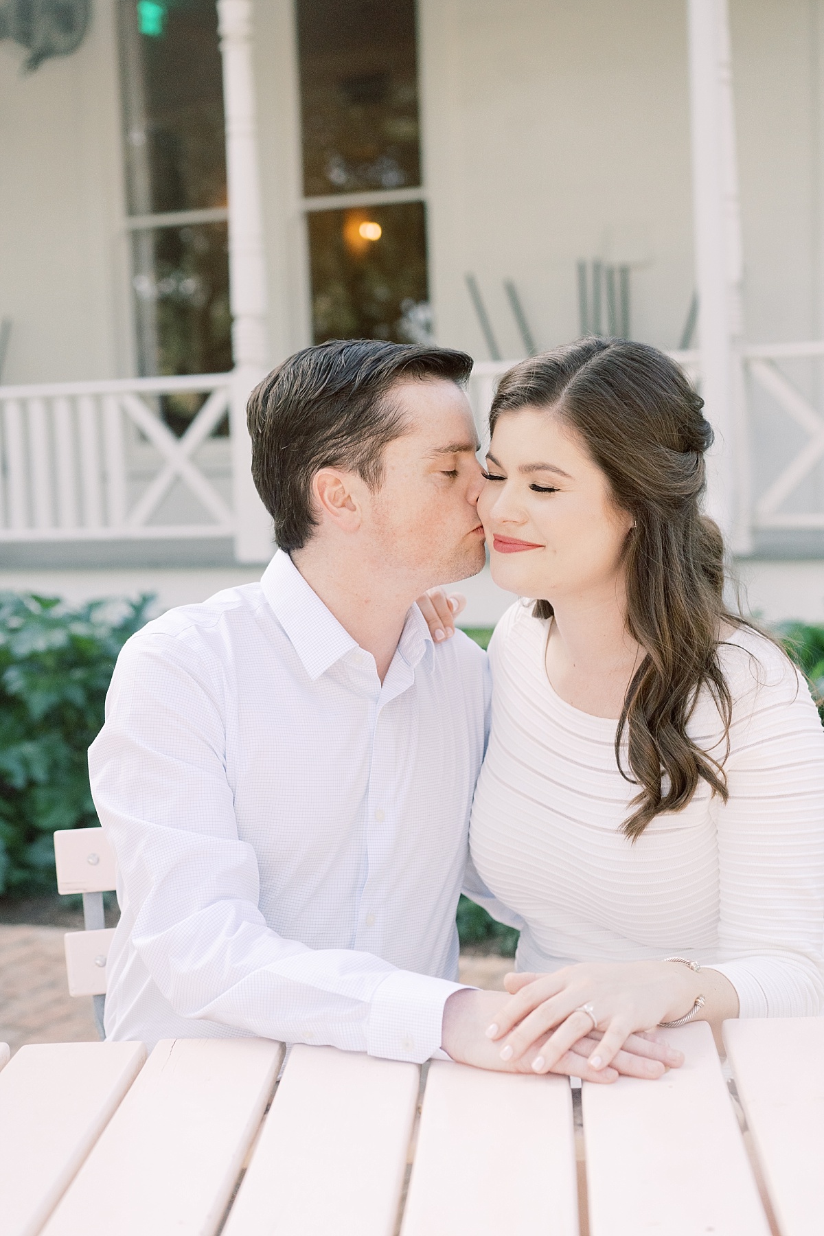 Engagement Session at Mattie's, A beautiful woman in a white dress smiles as her handsome fiance kisses her cheek in front of a southern porch.