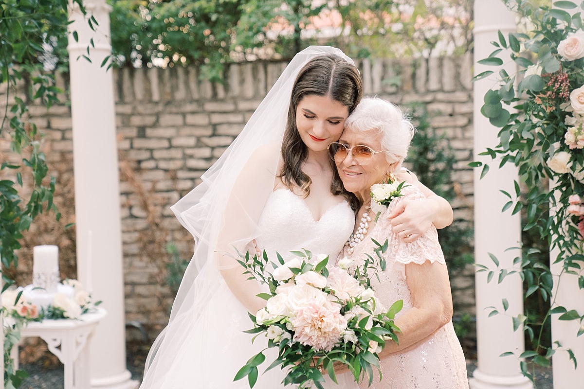 Brunette bride wraps her arm around her grandmother while standing under a beautiful archway for wedding photography with Paige Vaughn Photography in Austin, Texas.