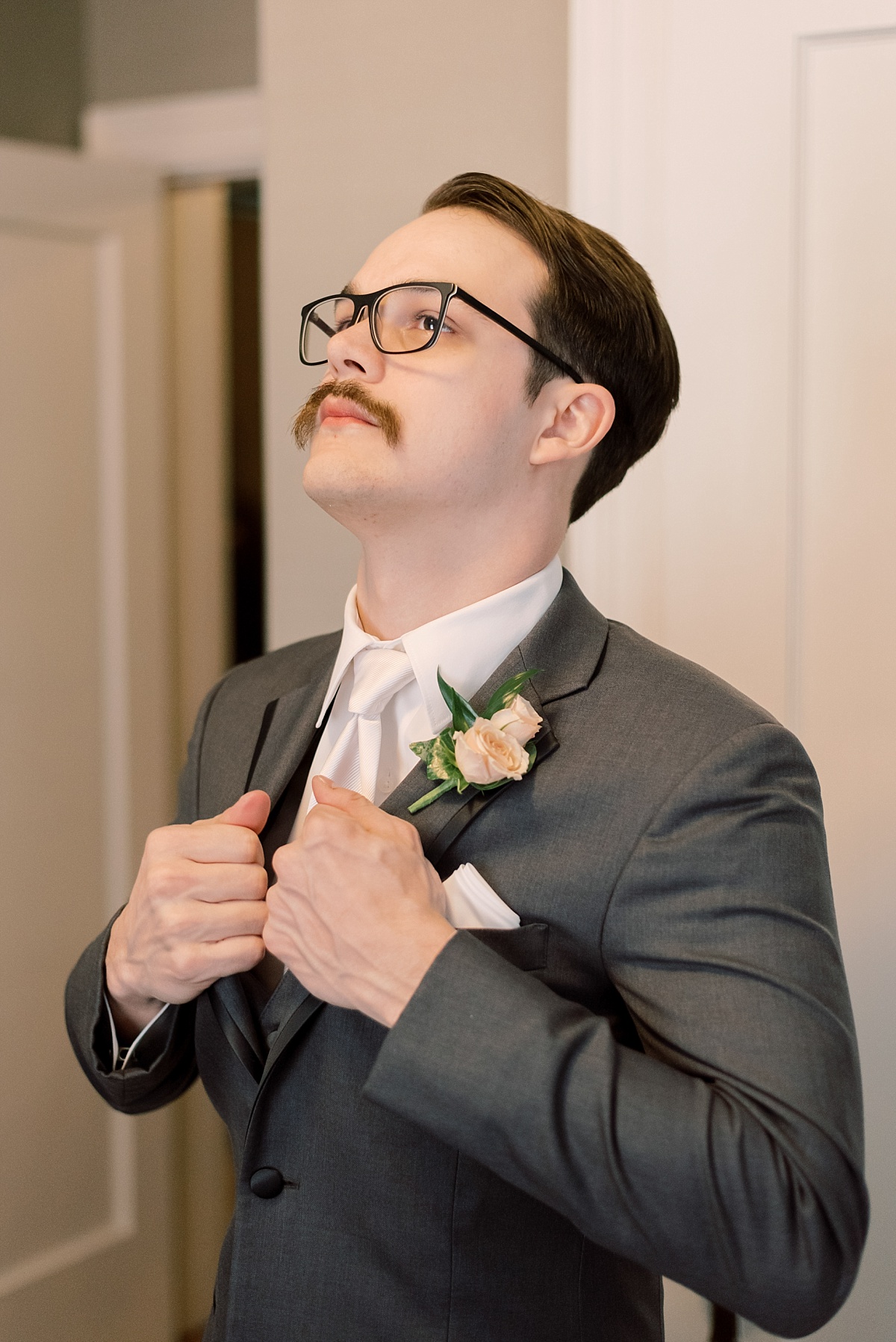Groom with glasses and a mustache prepares for his wedding day in gray tux while having his wedding photographs taken by Paige Vaughn Photography in Austin, Texas.