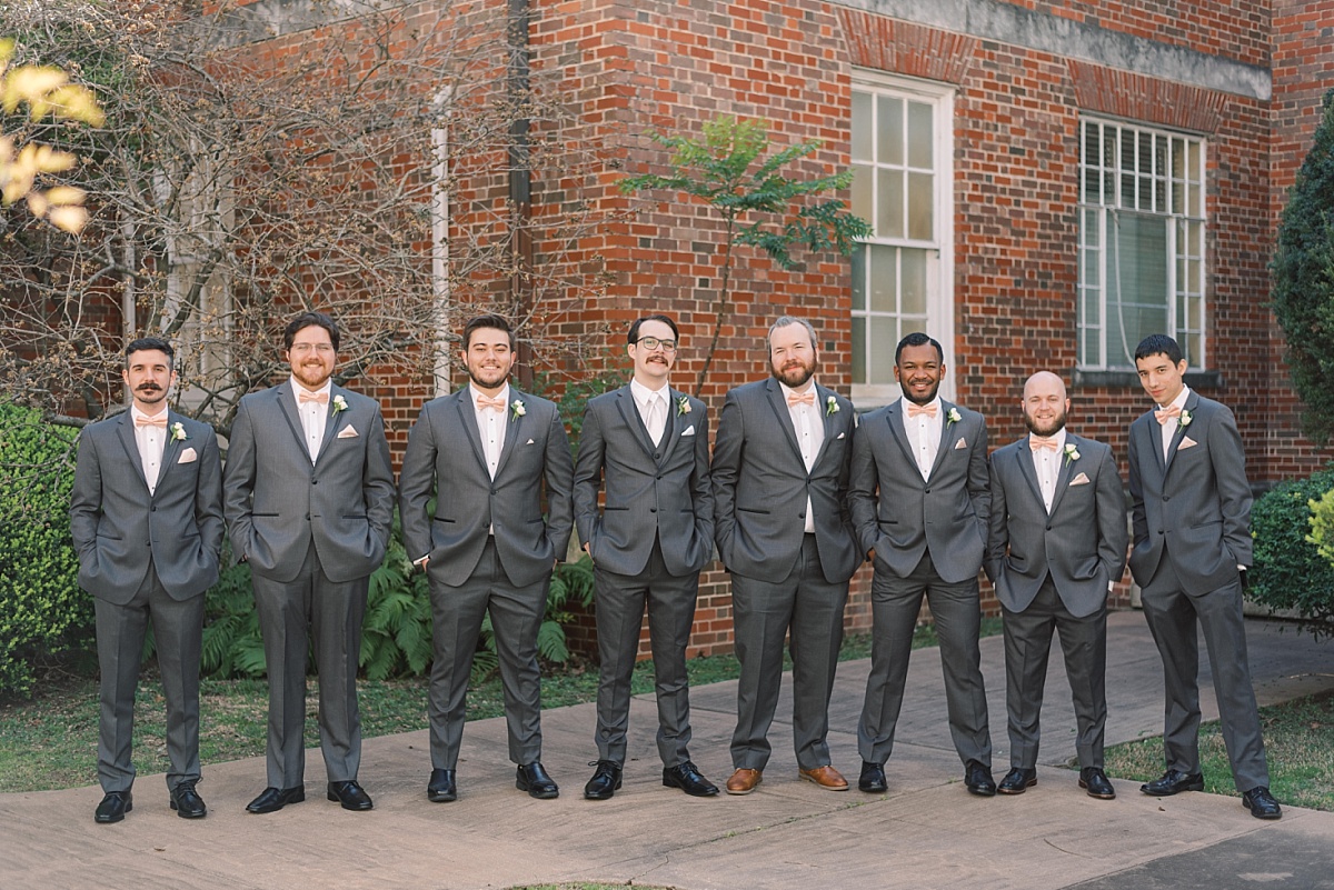 A Caucasian groom stands with his seven-person wedding party, wearing gray tuxes and pink bow ties while posing for wedding photographs with Paige Vaughn in Austin, Texas.