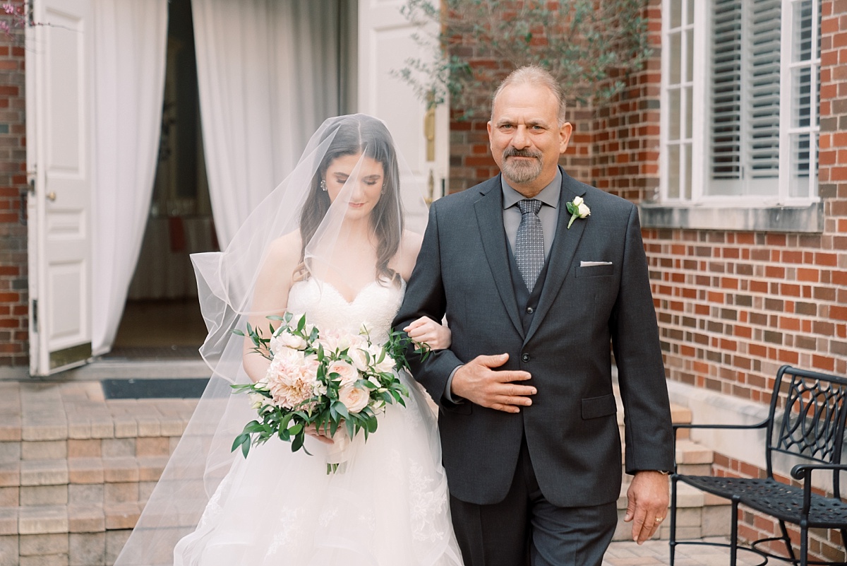Beautiful brunette bride in a white dress and veil walks arm in arm down the aisle with her father while having wedding photos taken by Paige Vaughn Photography at The Mansion in Austin, Texas.