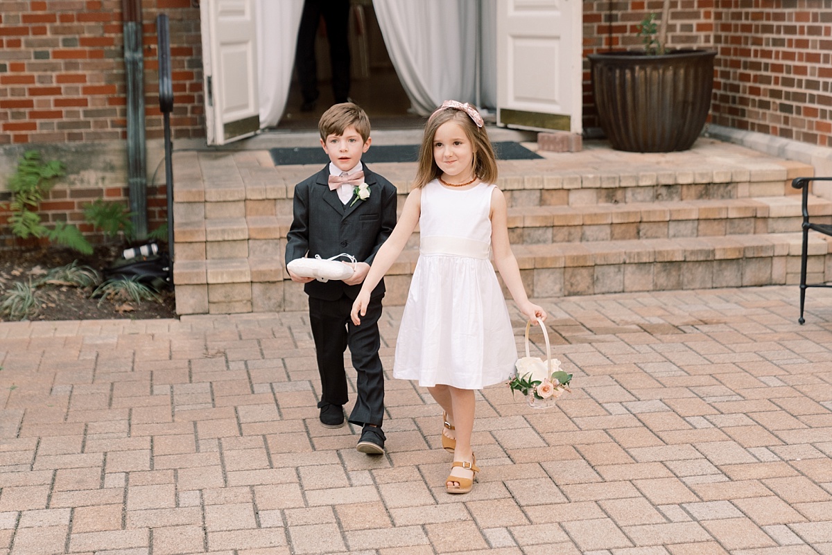 Adorable young flower girl in a white dress holding a basket and ring bearer in a gray tux approach the aisle while having photos taken by a wedding photographer in Austin, Texas.