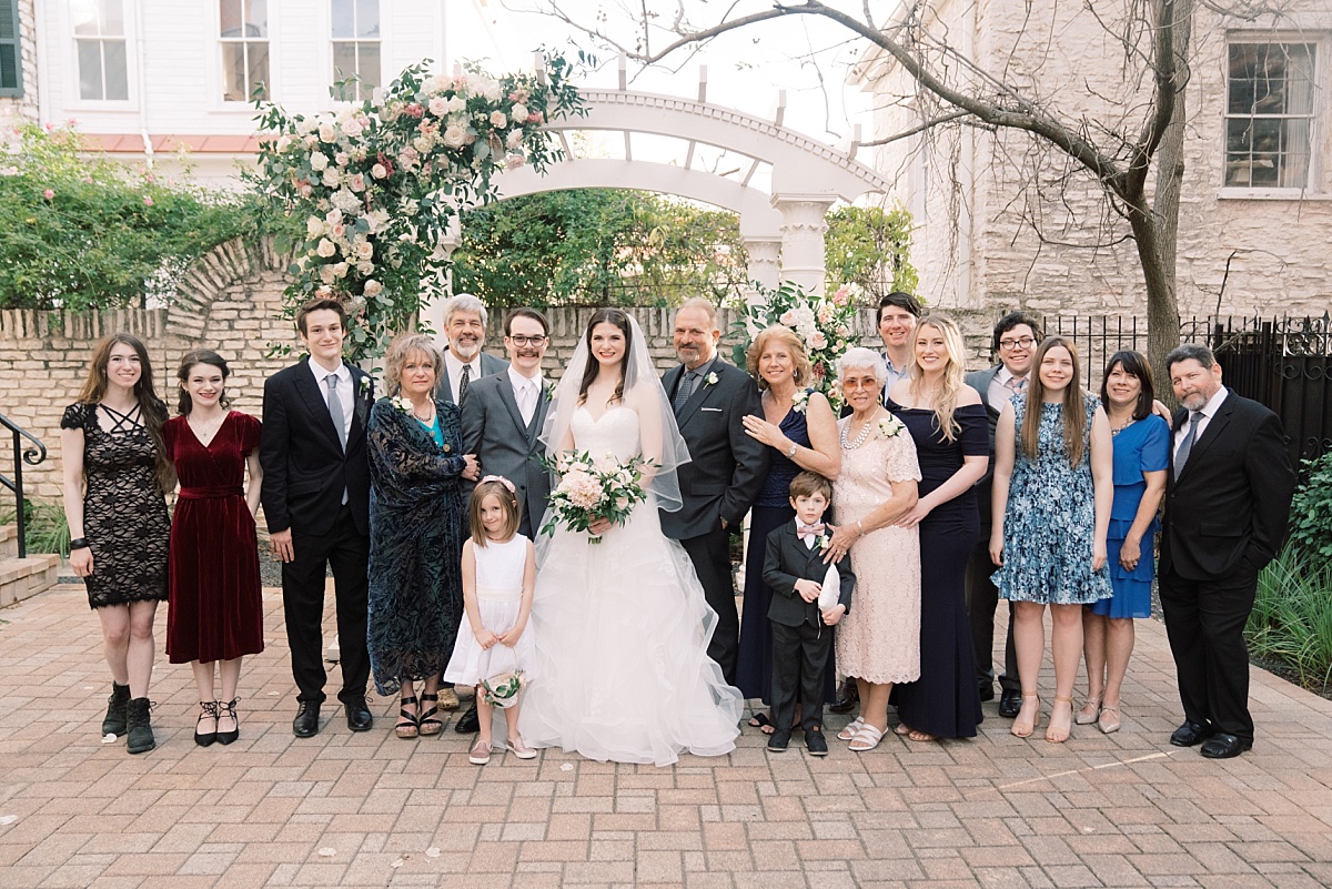 A bride and groom stand with family in front of a beautiful white alter at The Mansion, Austin, Texas while posing for wedding photos with Paige Vaughn Photography.