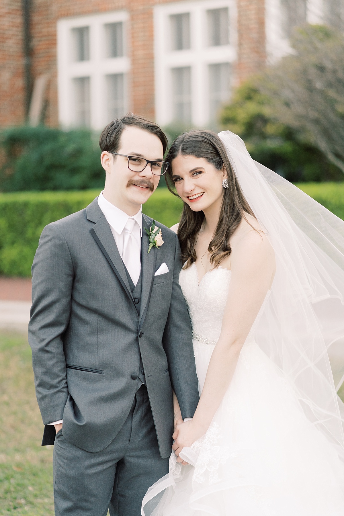 A newlywed Caucasian bride and groom stand together while holding hands and smiling at the camera for their wedding photographer in Austin, Texas.