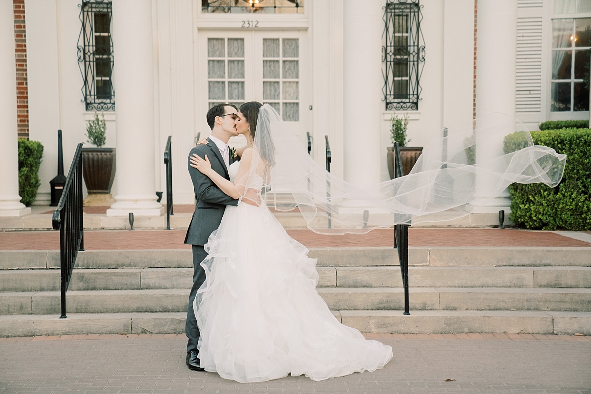 A southern bride and groom kiss while the bride's veil floats in the air in front of The Mansion in Austin, Texas.