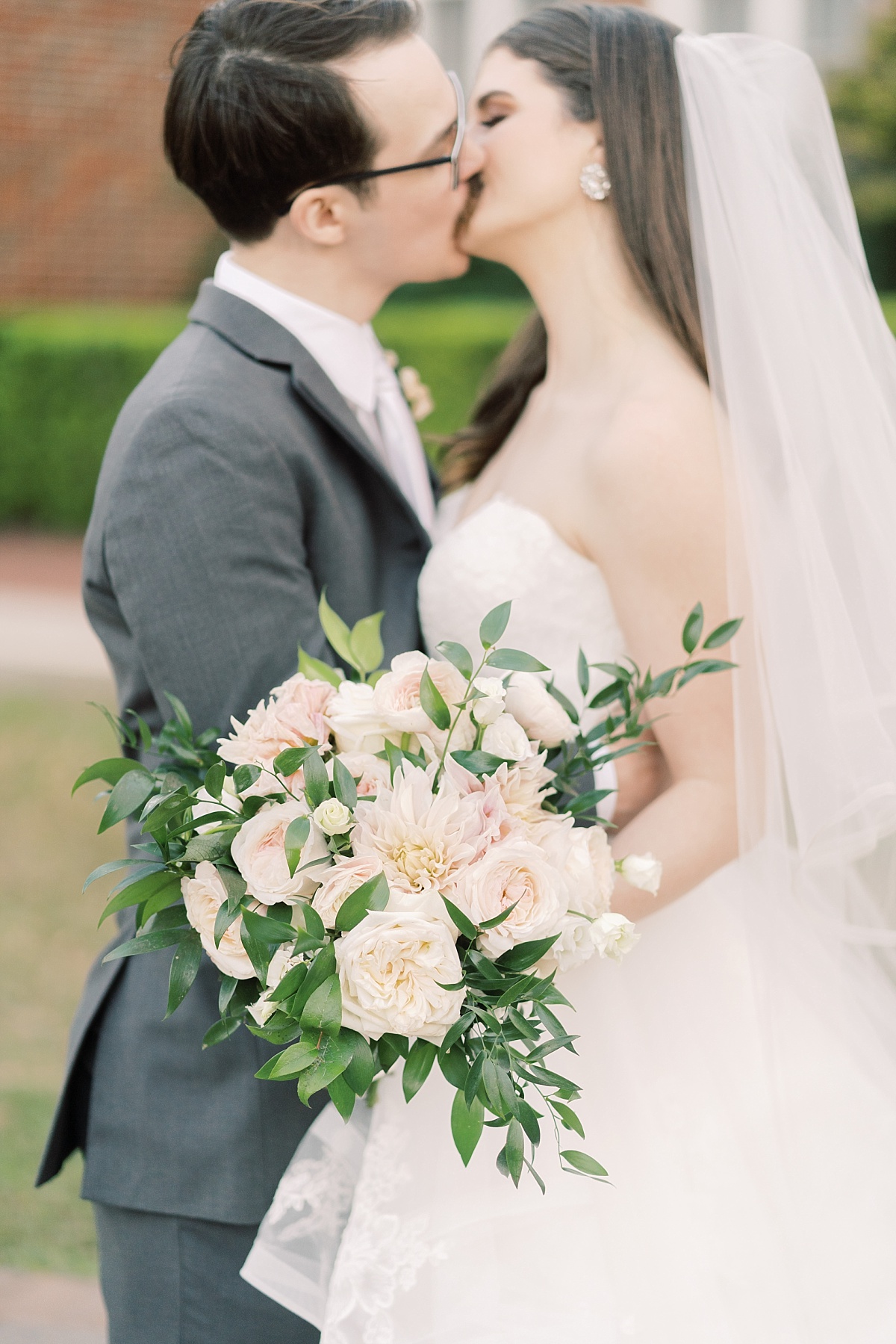 The camera focuses on a beautiful blush pink bouquet being held by a bride while kissing her new husband and having wedding photos taken by Paige Vaughn Photography.