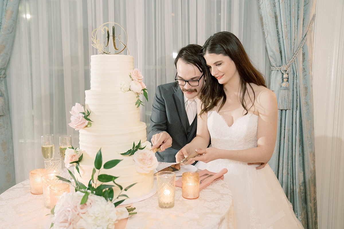 A wedding photographer in Austin, Texas captures a moment of a bride and groom slicing a white wedding cake decorated with pink flowers by Sweet Treets Bakery.