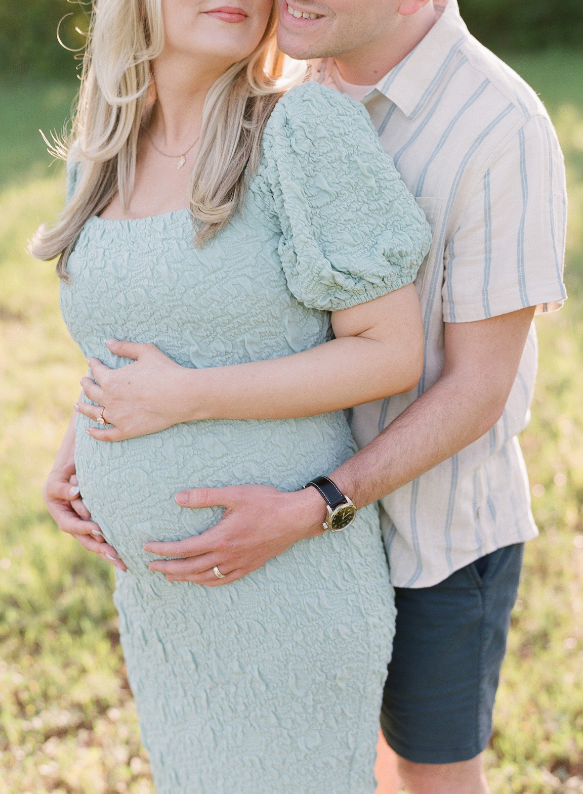 Golden hour film maternity photo of a couple embracing outdoors with soft sunlight filtering through the trees