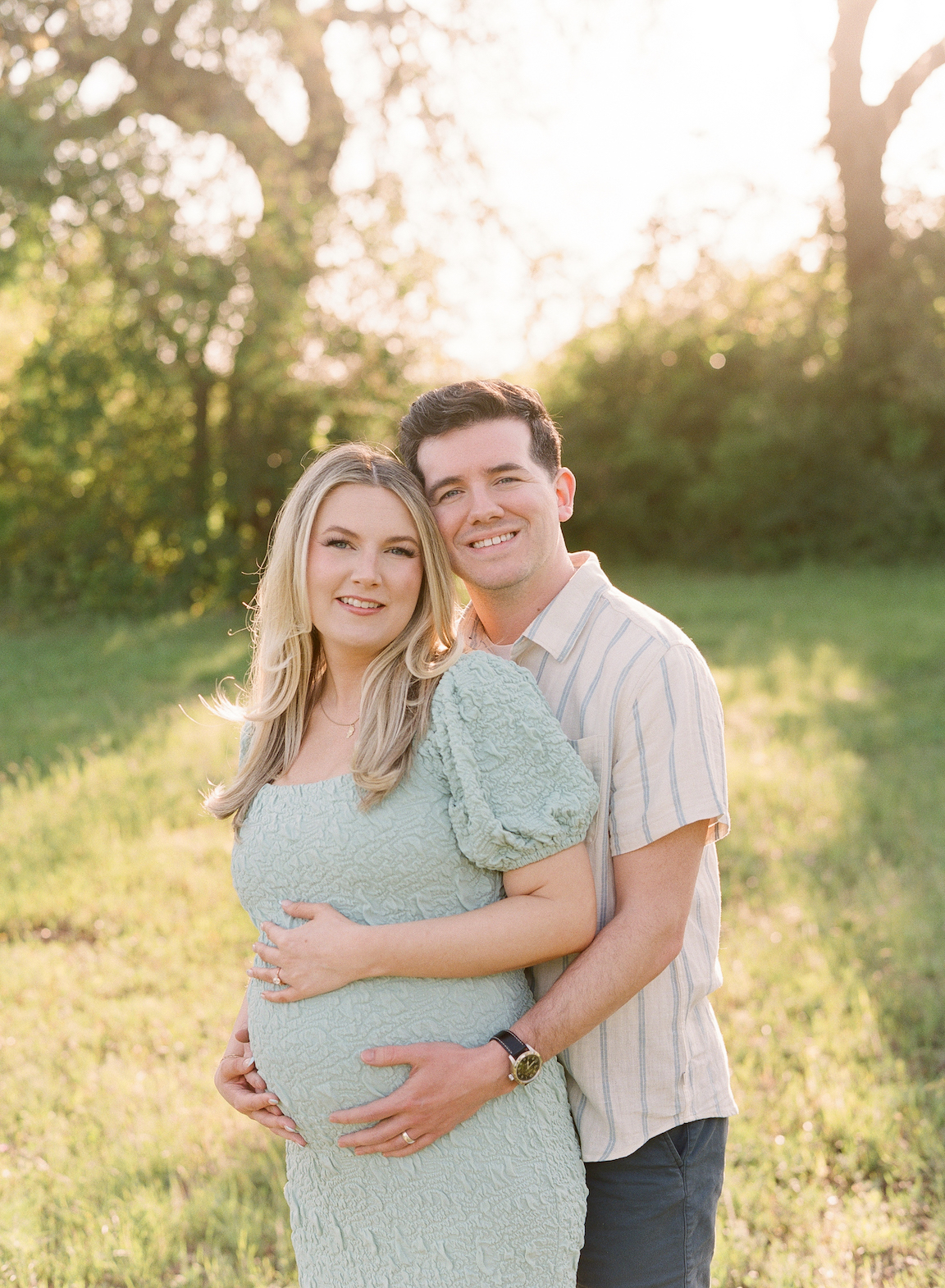 Golden hour film maternity photo of a couple embracing outdoors with soft sunlight filtering through the trees