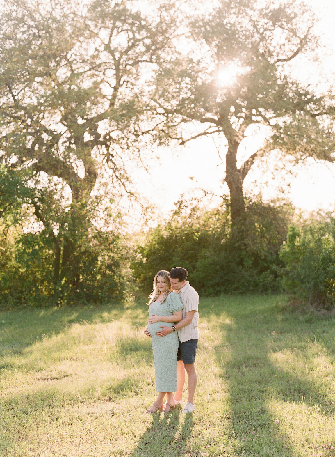 Golden hour film maternity photo of a couple embracing outdoors with soft sunlight filtering through the trees