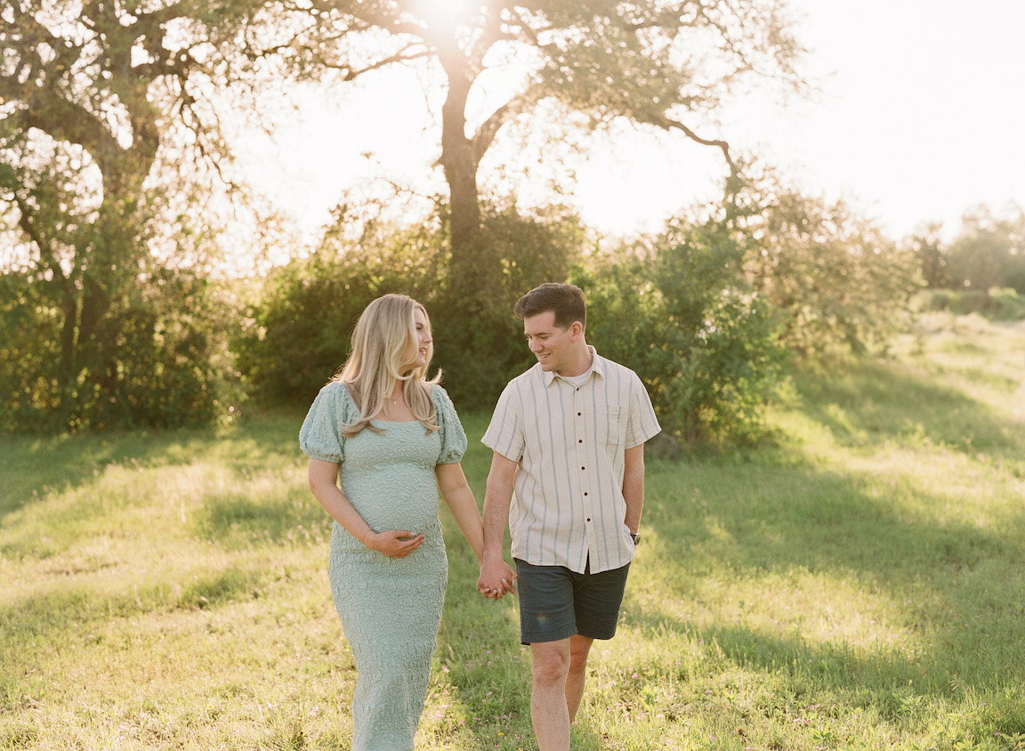 Golden hour film maternity photo of a couple embracing outdoors with soft sunlight filtering through the trees