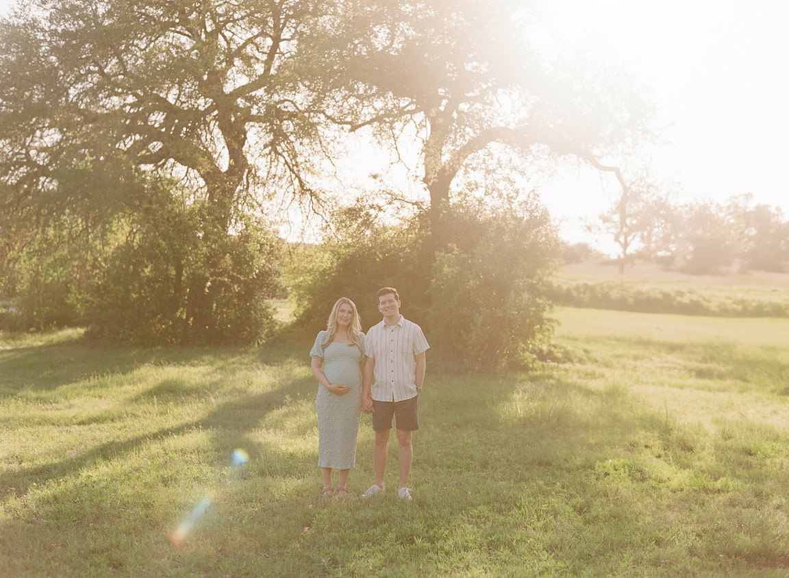 Golden hour film maternity photo of a couple embracing outdoors with soft sunlight filtering through the trees