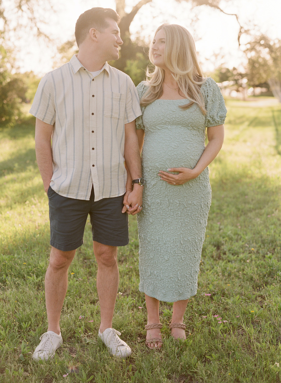 Golden hour film maternity photo of a couple embracing outdoors with soft sunlight filtering through the trees